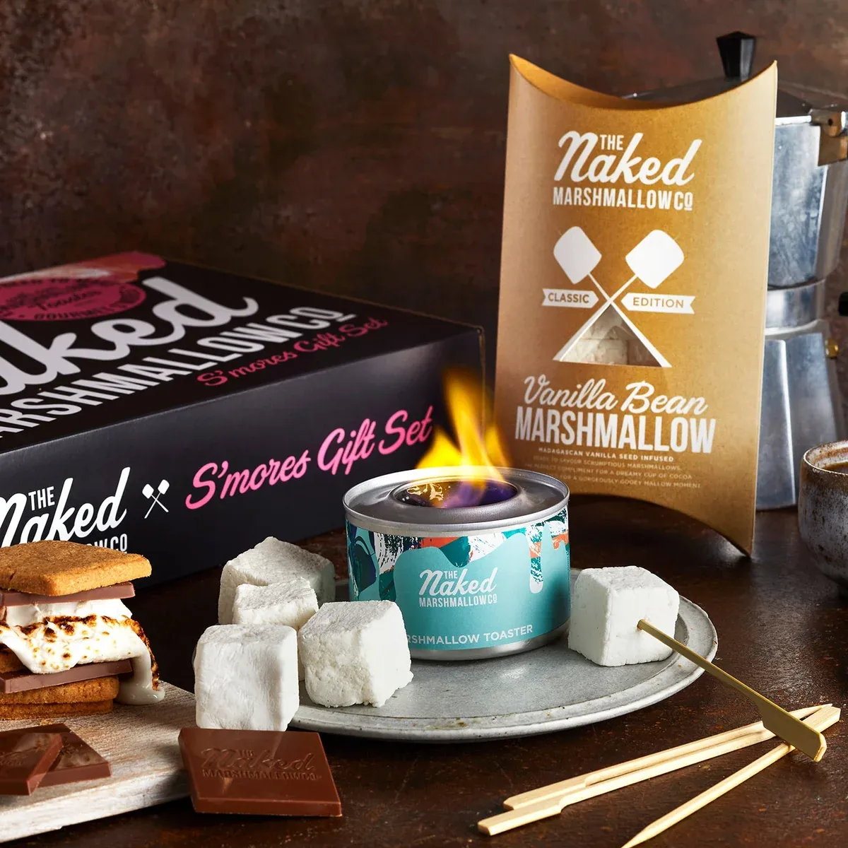 The Naked Marshmallow Co products styled on a dark wooden table: a black S'mores Gift Set box, a gold Vanilla Bean Marshmallow pillow pack, a lit teal marshmallow toaster surrounded by white marshmallow cubes, and a s'more with melting chocolate.